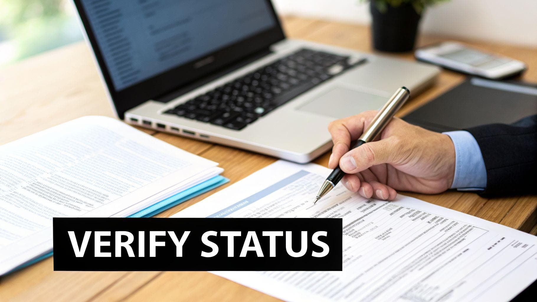 A person in a suit verifying status on documents with a pen, laptop, and phone on a wooden desk.